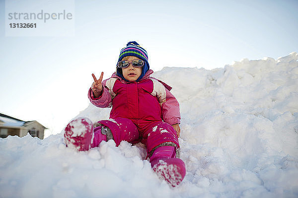 Mädchen mit Sonnenbrille zeigt Friedenszeichen  während sie auf Schnee vor klarem Himmel sitzt