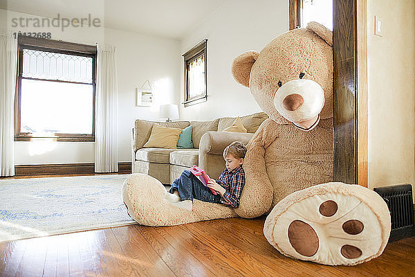 Seitenansicht eines Jungen  der zu Hause auf einem großen Teddybären sitzt und ein Spiel spielt