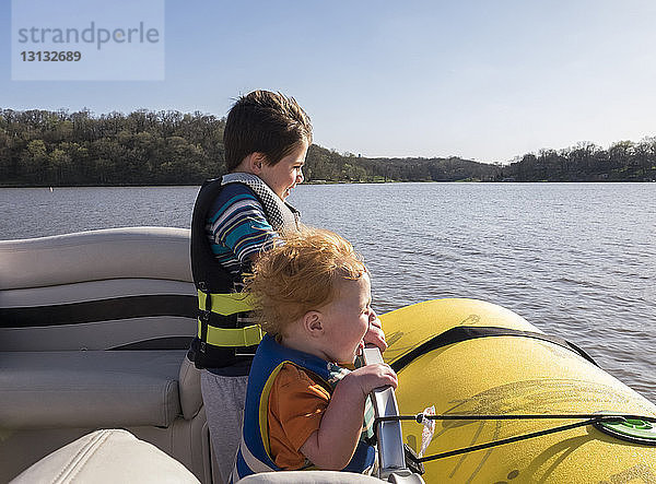 Glückliche Brüder tragen Schwimmwesten  während sie bei strahlendem Sonnenschein im Boot auf dem See vor klarem Himmel stehen