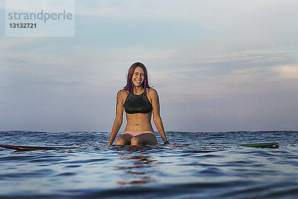 Glückliche Frau sitzt bei Sonnenuntergang auf einem Surfbrett im Meer