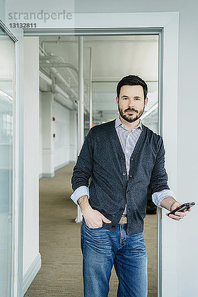 Porträt eines Geschäftsmannes  der ein Telefon hält  während er im Büro an der Tür steht