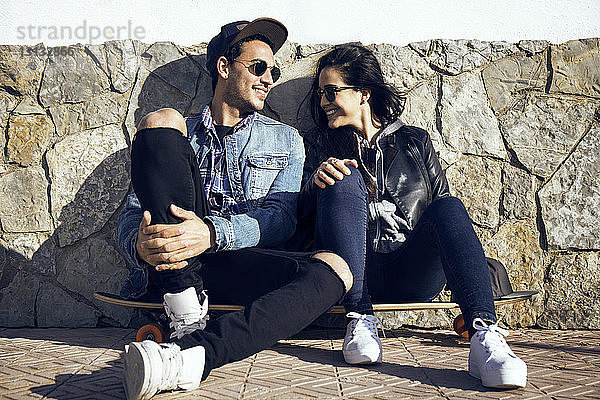 Glückliches Paar unterhält sich  während es auf einem Skateboard an einer Steinmauer sitzt