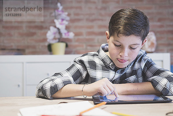 Junge streckt beim Zeichnen am Tablet-Computer bei Tisch die Zunge heraus