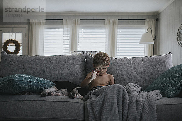 Junge ohne Hemd benutzt Tablet-Computer  während er zu Hause mit seinem Hund auf dem Sofa sitzt