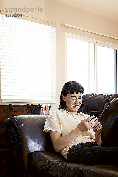 Lächelnde Frau telefoniert  während sie sich zu Hause auf dem Sofa entspannt