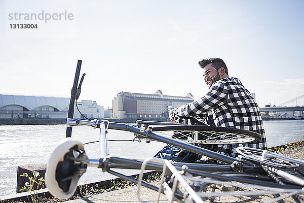 Junger Mann sitzt mit dem Fahrrad vor dem Kanal vor klarem Himmel