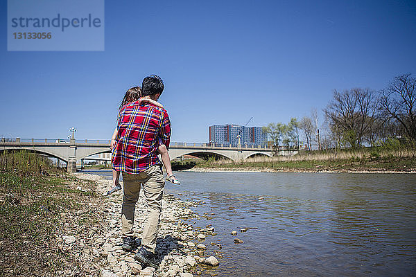 Vater trägt Tochter in voller Länge  während er am Flussufer gegen Brücke und klaren blauen Himmel am sonnigen Tag läuft