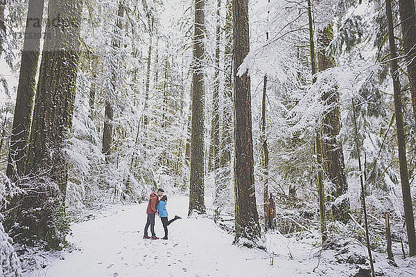 Paar küsst sich in voller Länge  während es im Winter im Lynn Canyon Park zwischen Bäumen im Wald steht