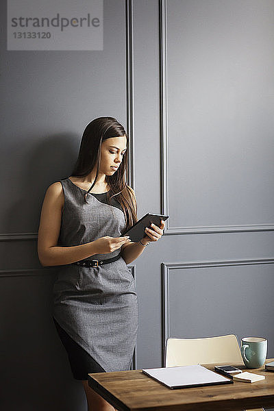 Geschäftsfrau benutzt Tablet-Computer  während sie sich im Büro an die Wand lehnt