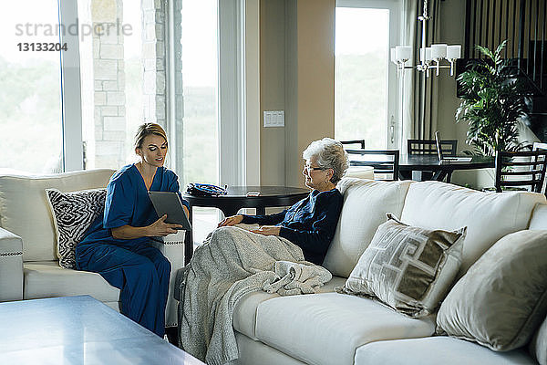 Heimpflegerin zeigt einer älteren Frau einen Tablet-Computer  während sie auf dem Sofa im Wohnzimmer sitzt