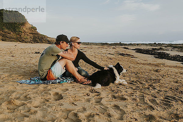 Paar mit Hund sitzt am Strand auf Sand