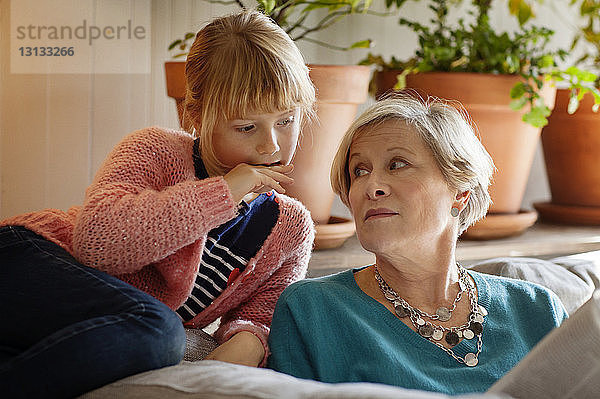 Großmutter betrachtet Enkelin beim Lesen einer Zeitschrift  während sie auf dem Sofa sitzt