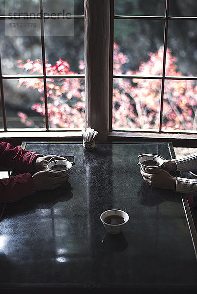 Ausgeschnittenes Bild eines Paares  das Kräuterteetassen bei Tisch durch ein Fenster hält