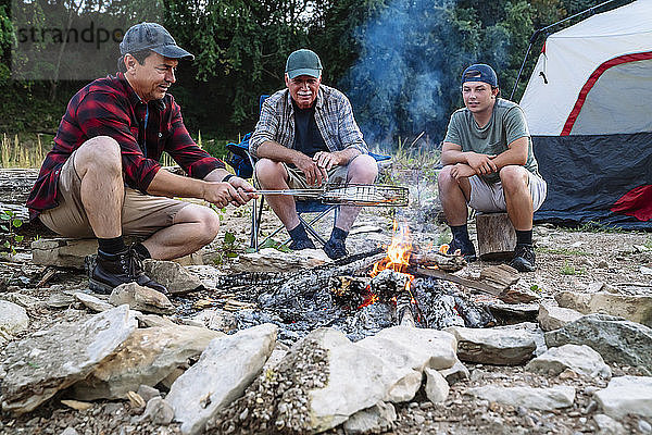 Mittelgroßer Mann kocht Fisch am Lagerfeuer  während er bei Sonnenuntergang mit Freunden auf dem Campingplatz sitzt
