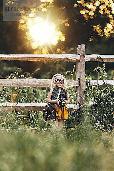Fröhliches Mädchen mit Kamera steht bei Sonnenuntergang auf Grasfeld vor Zaun