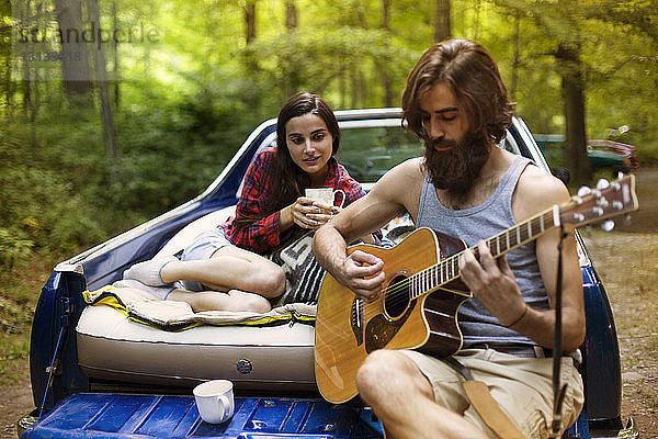 Mann spielt für seine Freundin Gitarre  während er im Pickup im Wald sitzt