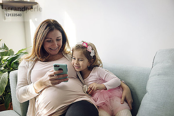 Mutter und Tochter schauen auf ein Smartphone  während sie auf dem Sofa an der Wand sitzen