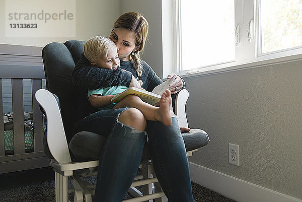 Mutter küsst Sohn beim Lesen eines Buches auf einem Stuhl zu Hause