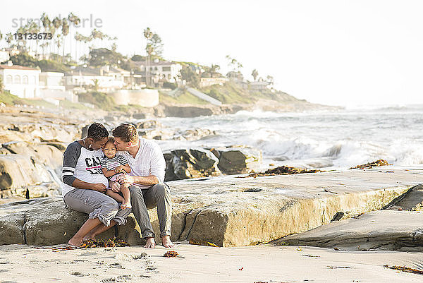 Eltern küssen ihre Tochter  während sie am Strand vor klarem Himmel sitzen