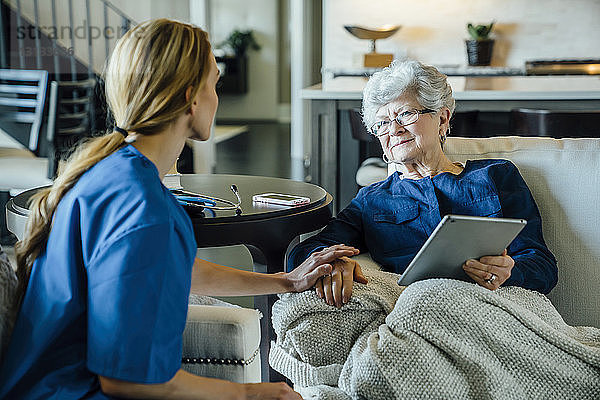 Ältere Frau schaut zur häuslichen Pflegekraft  während sie einen Tablet-Computer im Wohnzimmer hält