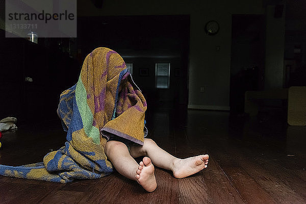 Verspielter kleiner Junge  der den Kopf mit einem Handtuch bedeckt  während er zu Hause auf dem Boden sitzt