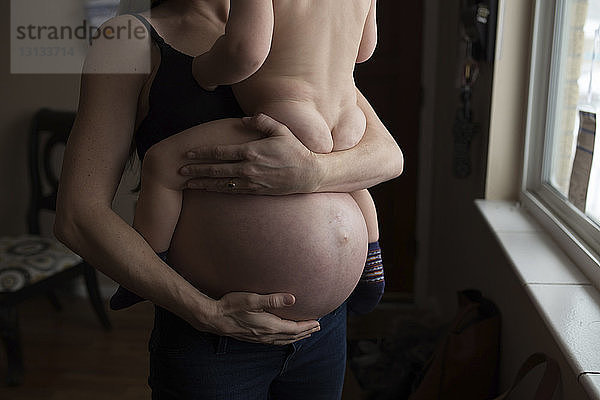 Mittelsektion einer schwangeren Mutter  die einen Sohn trägt  während sie zu Hause ihren Bauch gegen das Fenster streicht