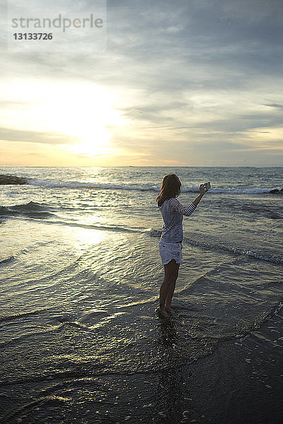 Frau hält Mobiltelefon in der Hand  während sie bei Sonnenuntergang am Strand vor bewölktem Himmel steht