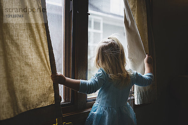 Rückansicht eines zu Hause stehenden Mädchens  das durch ein Fenster schaut