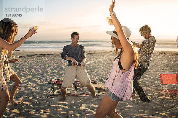 Glücklicher Mann öffnet Weinflasche  während Freunde am Strand tanzen