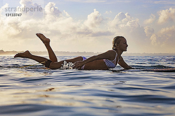 Seitenansicht einer Frau  die auf einem Surfbrett im Meer vor bewölktem Himmel liegt