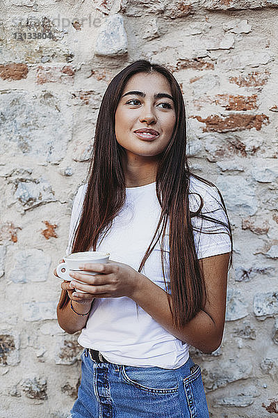 Frau hält Cappuccino in der Hand und schaut weg  während sie an alter Ziegelmauer steht