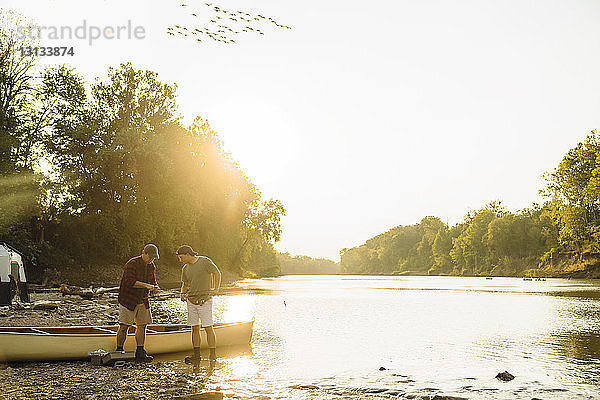 Freunde stellen die Angelrute ein  während sie bei Sonnenuntergang am Seeufer beim Boot stehen