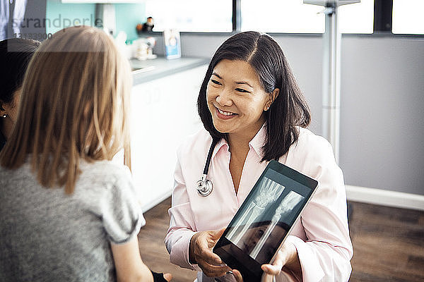 Lächelnde Ärztin zeigt einem Mädchen in der Klinik ein Hand-Röntgenbild am Tablet-Computer