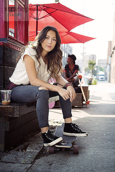 Porträt einer jungen Frau  die mit einem Skateboard auf einer Stützmauer sitzt