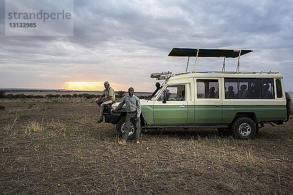 Porträt von Freunden im Geländewagen vor bewölktem Himmel im Serengeti-Nationalpark bei Sonnenuntergang