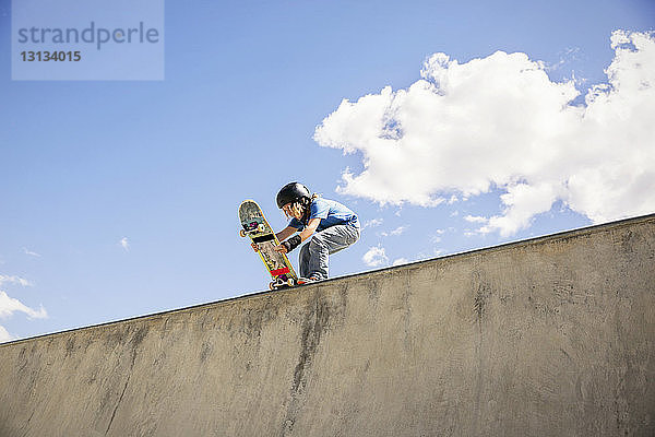 Niedrigwinkelansicht eines Jungen  der ein Skateboard auf einer Rampe vor blauem Himmel platziert