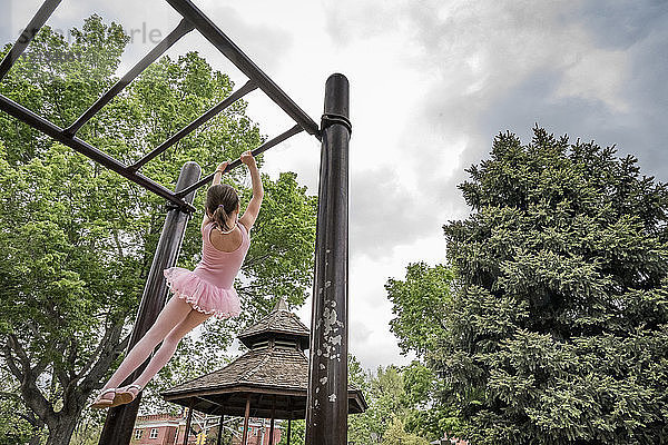Rückansicht eines Mädchens in Ballettkostüm  das an Affenstangen vor bewölktem Himmel auf dem Spielplatz hängt