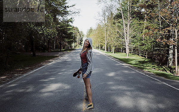 Ganzflächiges Porträt einer fröhlichen Frau mit Skateboard  die auf der Straße inmitten von Bäumen steht