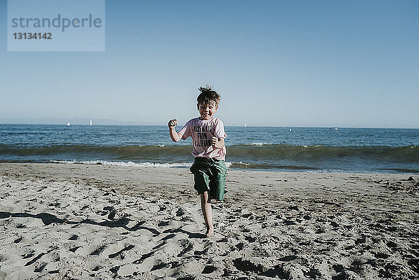 Sorgloser Junge rennt am Strand gegen den klaren Himmel am sonnigen Tag
