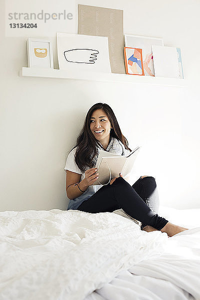 Glückliche Frau hält Tagebuch  während sie zu Hause auf dem Bett sitzt