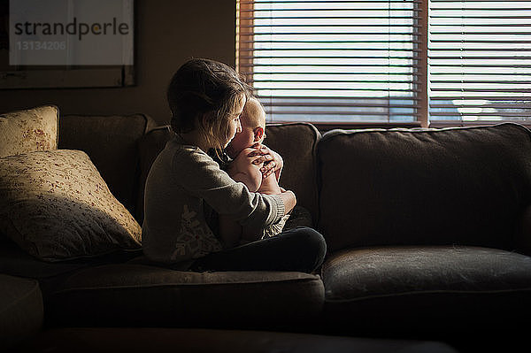 Seitenansicht eines Mädchens  das einen kleinen Jungen umarmt  während es zu Hause auf dem Sofa sitzt