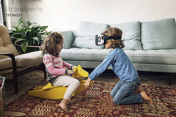 Junge trägt einen Virtual-Reality-Simulator und spielt mit seiner Schwester  die zu Hause auf einem Schaukelpferd sitzt