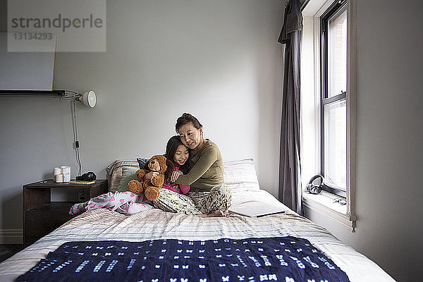 Mutter umarmt Tochter  während sie zu Hause auf dem Bett sitzt