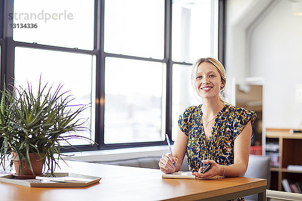 Porträt einer lächelnden Geschäftsfrau mit Mobiltelefon  die im Büro am Tisch sitzt