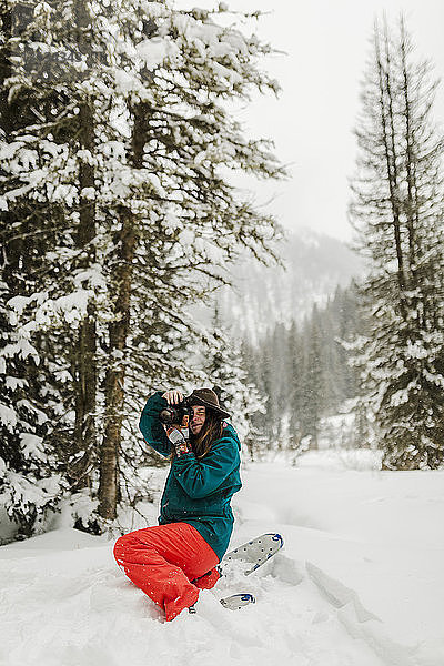 Frau fotografiert mit der Kamera  während sie auf einem schneebedeckten Feld im Wald kauert