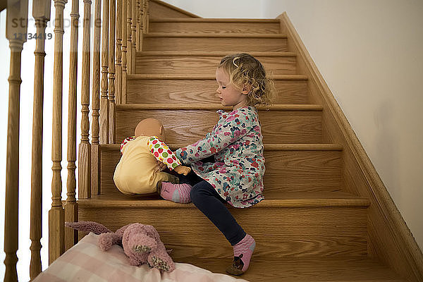 Seitenansicht eines Mädchens  das mit einer Puppe spielt  während es zu Hause auf Holzstufen sitzt