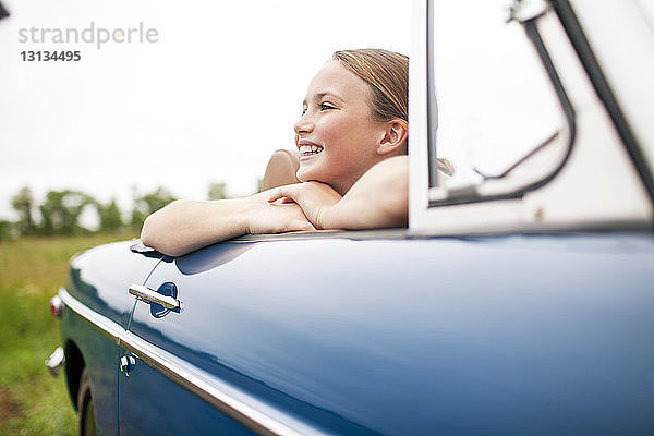 Glückliches Mädchen sitzt im Auto am Feld gegen den Himmel