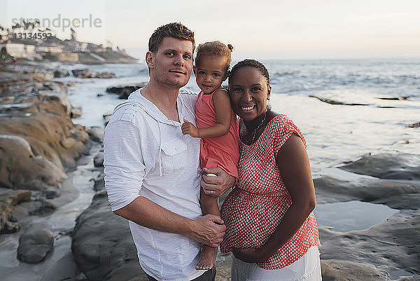 Porträt von glücklichen Eltern mit Tochter am Strand bei Sonnenuntergang