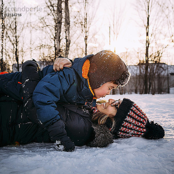 Glückliche Mutter mit Sohn liegt bei Sonnenuntergang auf schneebedecktem Feld gegen den Himmel