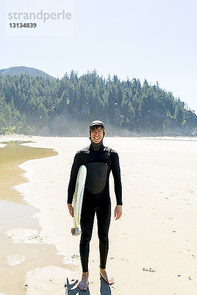 Porträt eines glücklichen Mannes  der ein Surfbrett hält  während er am Strand steht